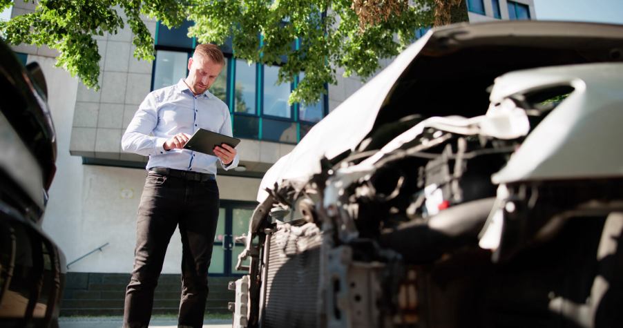 An insurance adjuster stands next to damaged car with a tablet