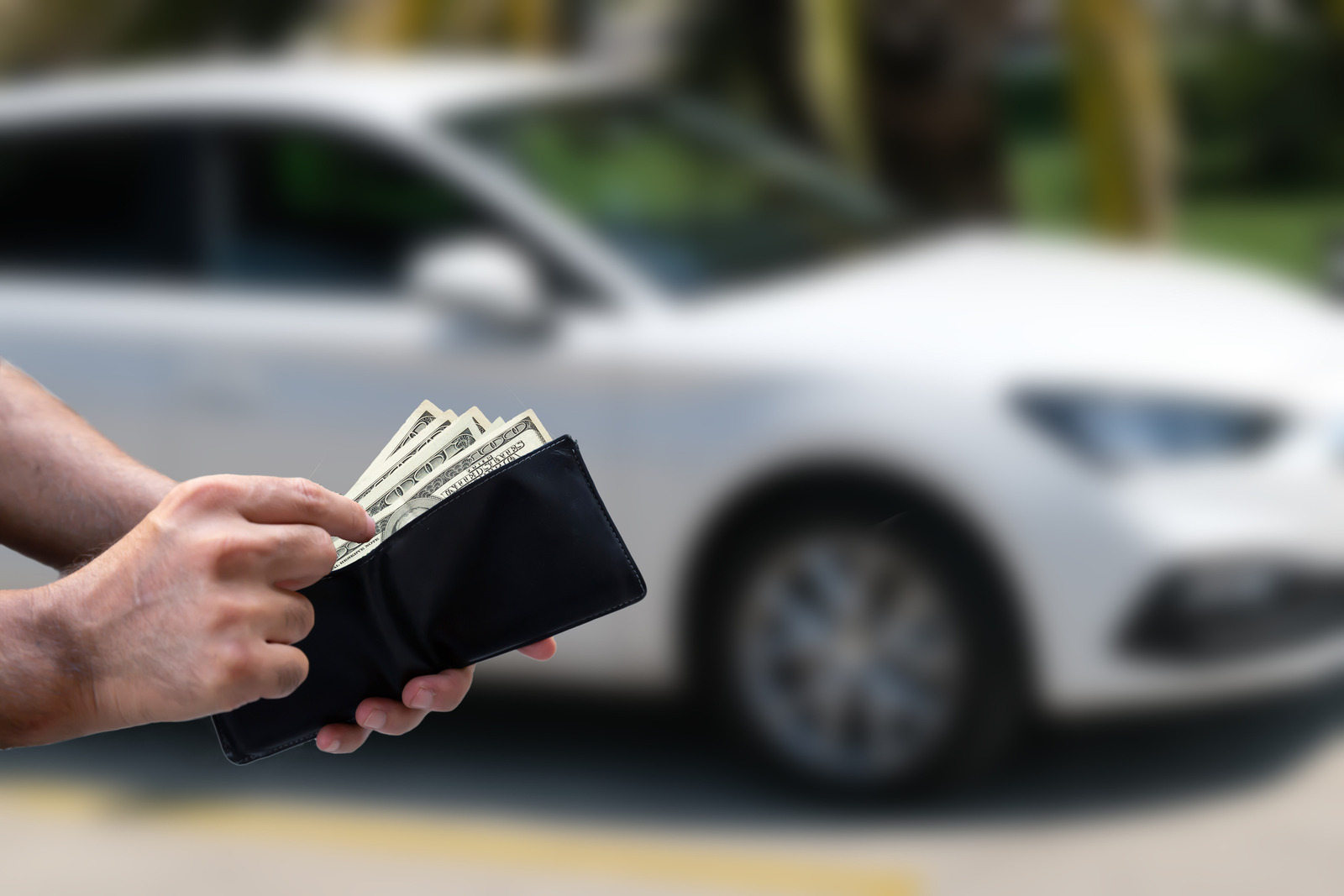 A man's hands holding a wallet with cash in it in front of a white sedan in background
