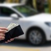 A man's hands holding a wallet with cash in it in front of a white sedan in background