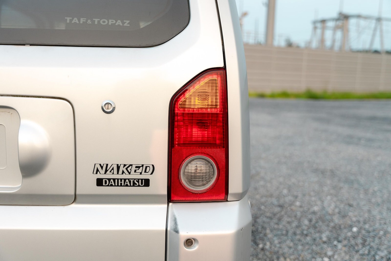 The "Naked" badge on the rear hatch of a Daihatsu Kei-car, a gravel lot visible in the background
