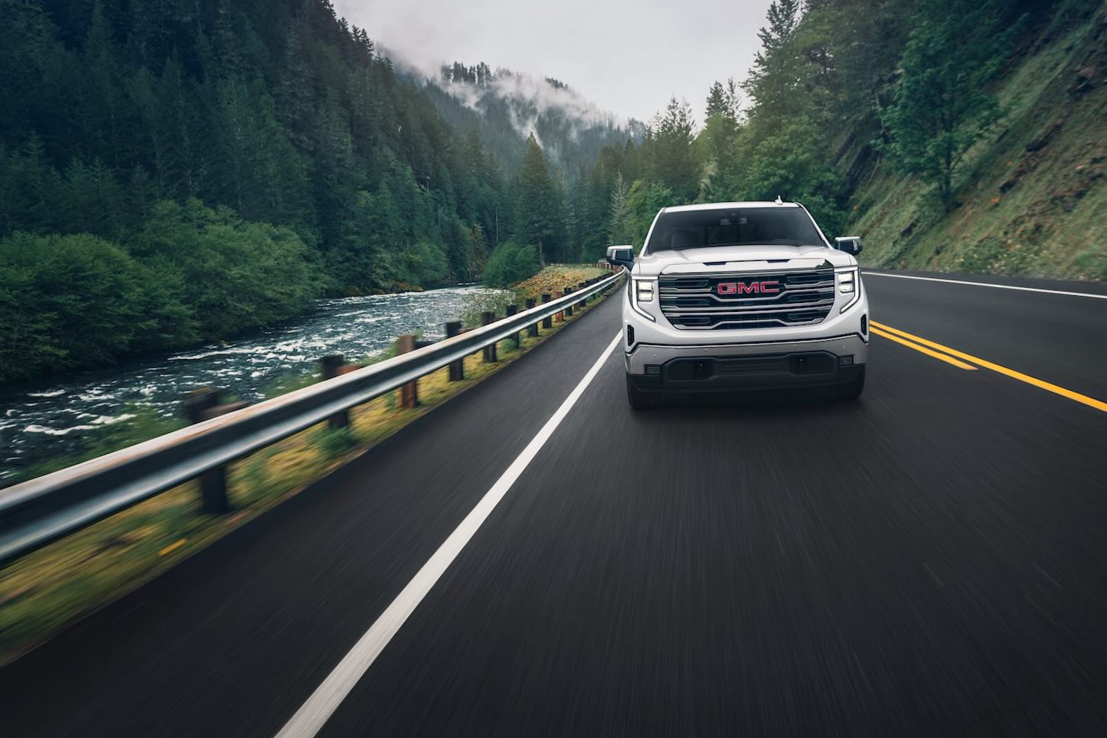 White GMC pickup truck drives along a road on a riverbank, mountains visible in the background.