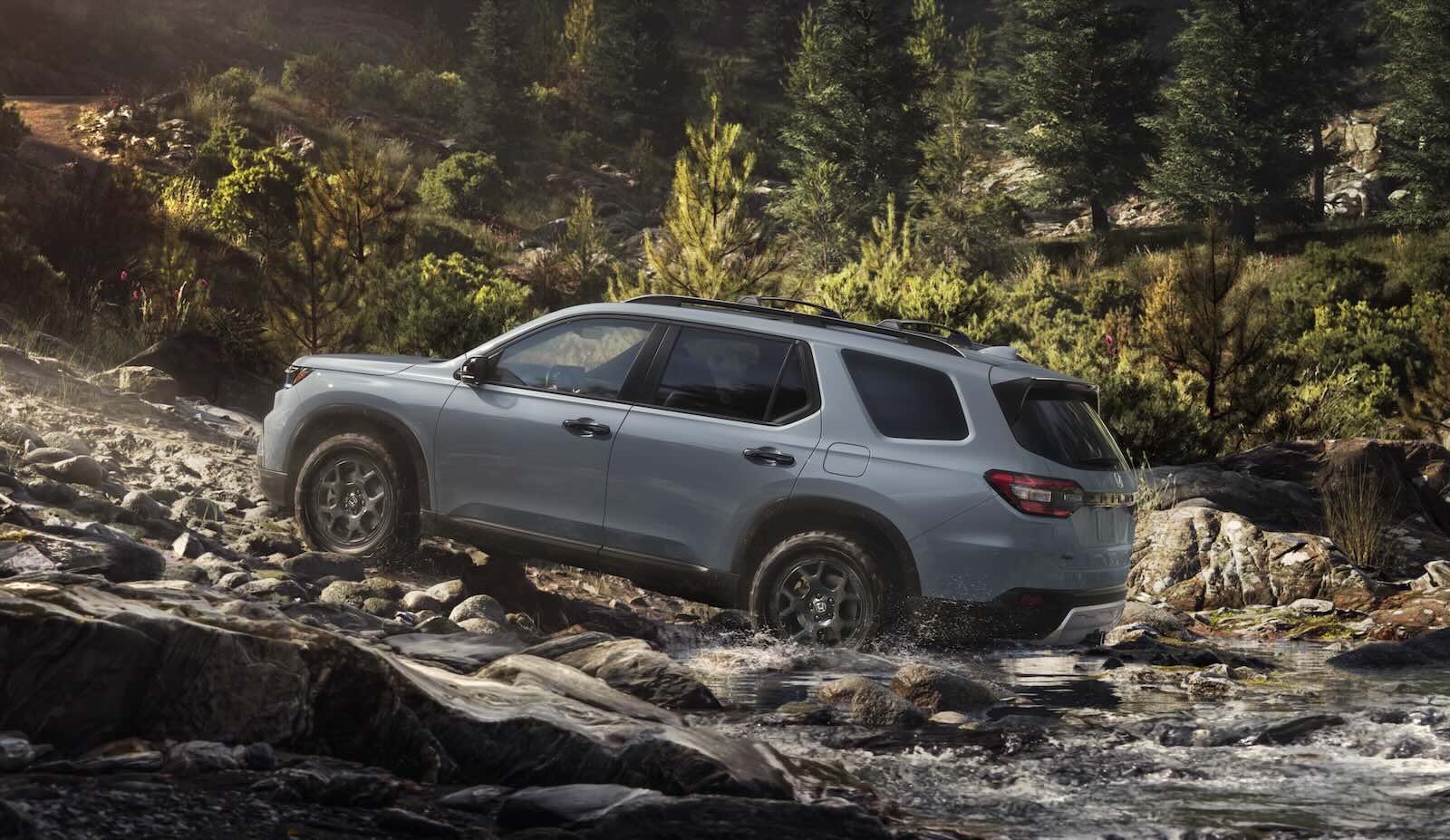 Honda Pilot SUV fording a river and driving up a gravel road, mountains visible in the background.