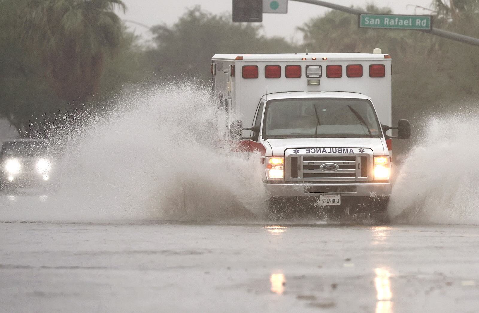 Ambulance races through standing water, road signs visible in the background.