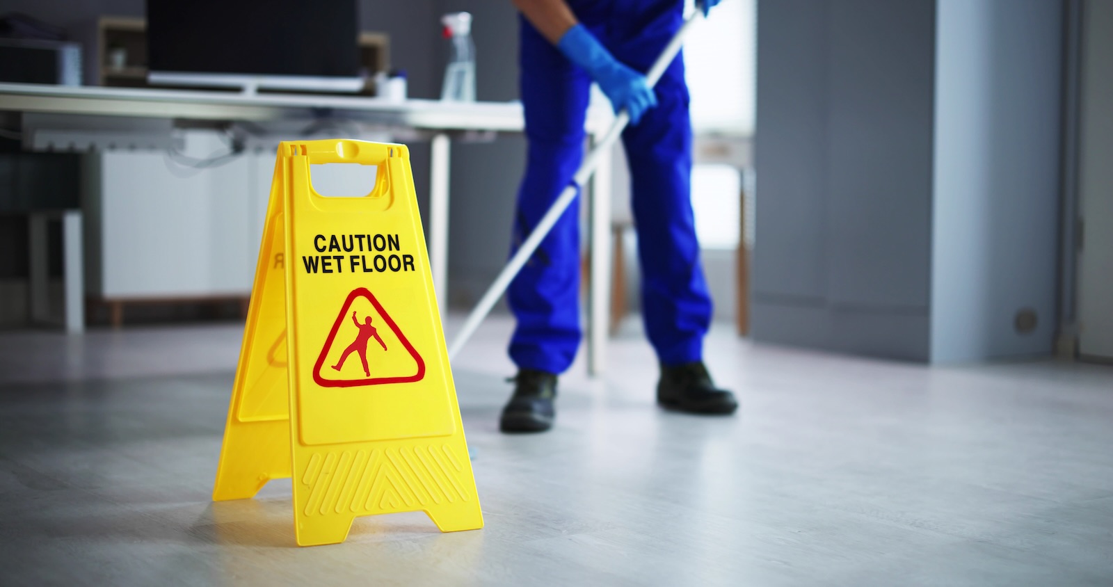 Lower half of a custodian mopping the floor in a high school, yellow caution sign in the foreground.