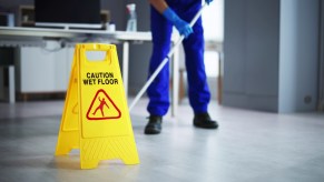 Lower half of a custodian mopping the floor in a high school, yellow caution sign in the foreground.