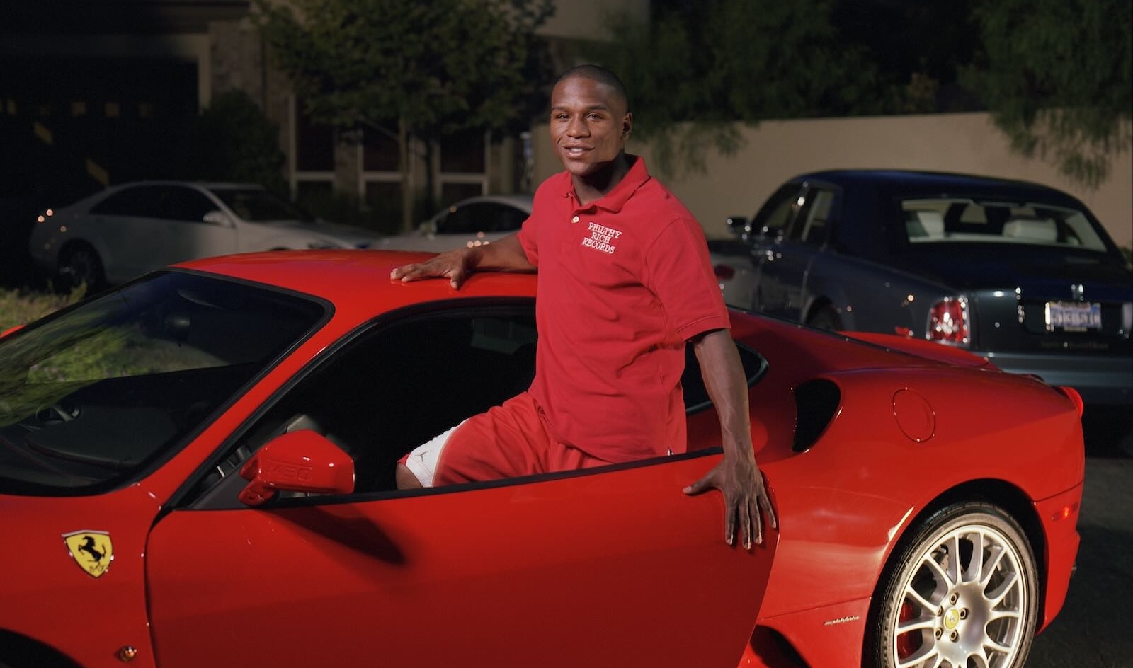 Floyd Mayweather Jr. smiles while standing in the open door of his red Ferrari, his driveway visible in the background.