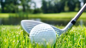 Golf ball and club on a course, pond visible in the background.