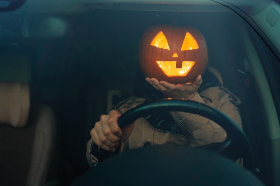 Car driver holding a jack o lantern pumpkin in front of their face while holding the steering wheel on Hallowen