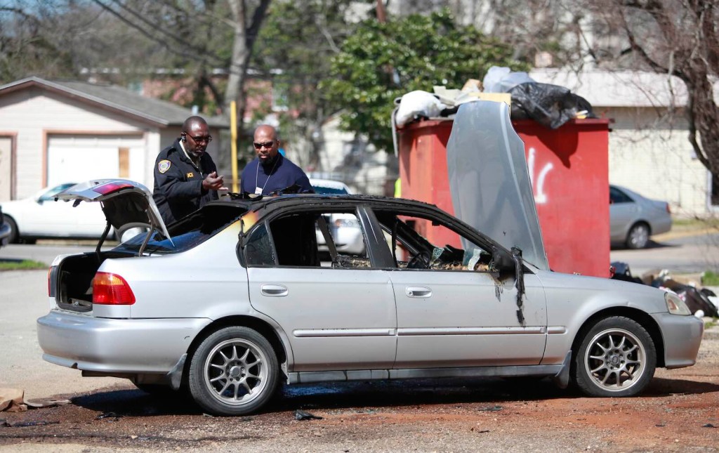 Police officers examine a Honda Civic that's been stolen, burned, and abandoned by a dumpster