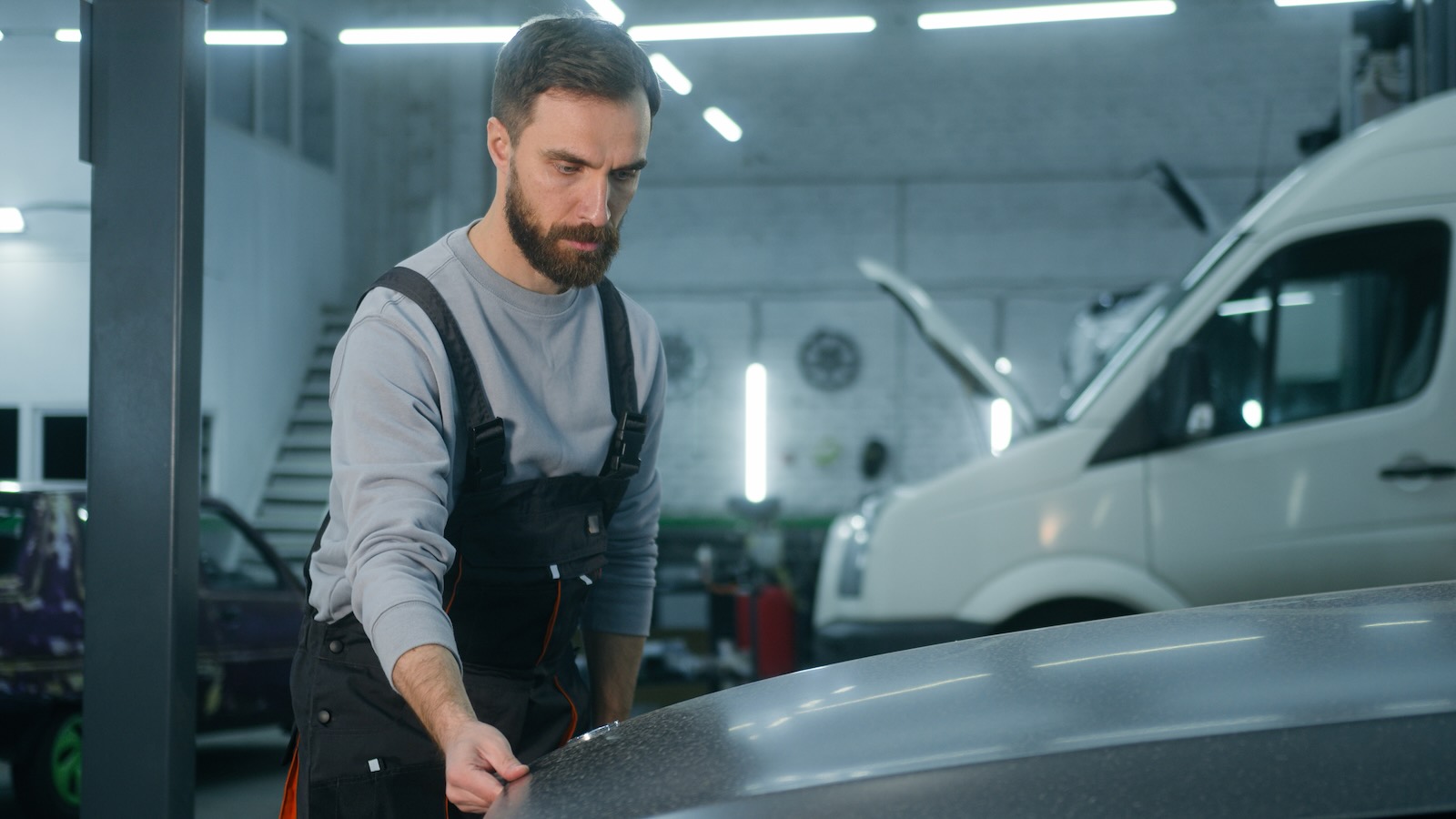 An auto mechanic holds the hood of a car while examining its logo badge, an auto shop visible in the background.