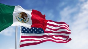 The flags of both the USA and Mexico waving in front of a blue sky