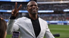 Actor Terry Crews wearing a white suit waves from a football field, the stands visible in the background.