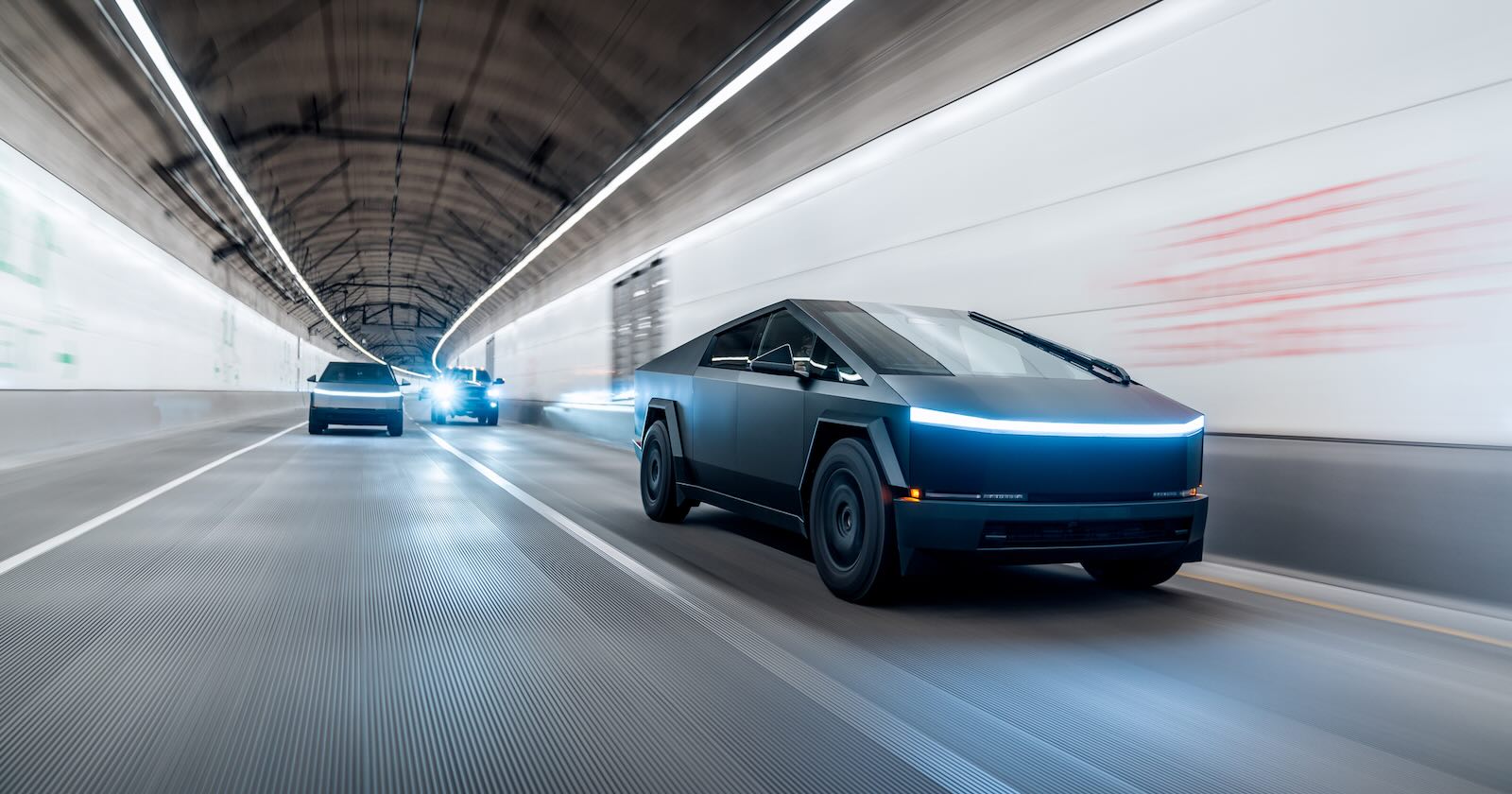 Black and silver Cybertruck driving through a tunnel at night, their headlights on.