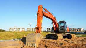 Tracked construction excavator on a job site, buildings visible in the background.