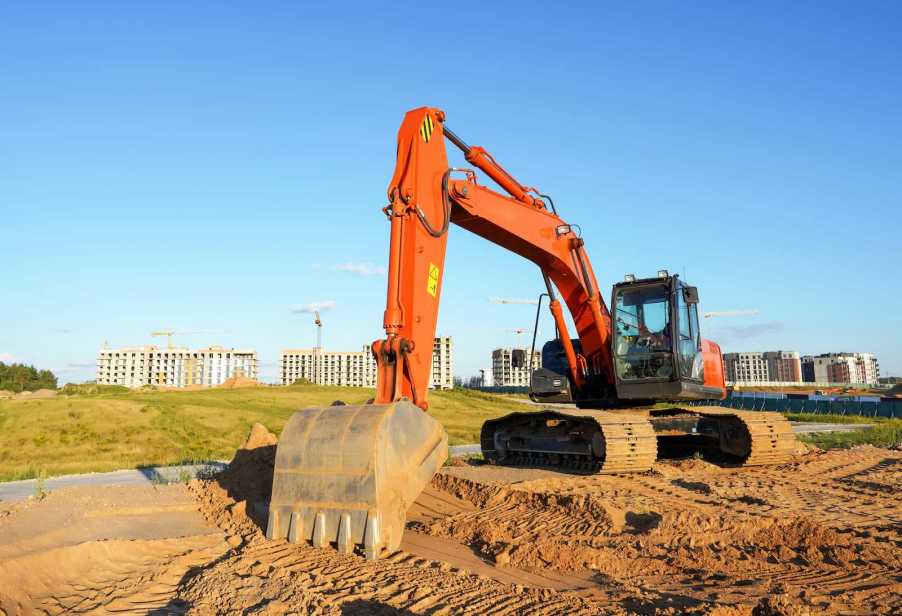 Tracked construction excavator on a job site, buildings visible in the background.