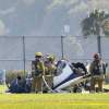 Fire fighters surround a small, ultralight airplane crash on a field, palm trees visible in the background.