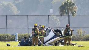 Fire fighters surround a small, ultralight airplane crash on a field, palm trees visible in the background.