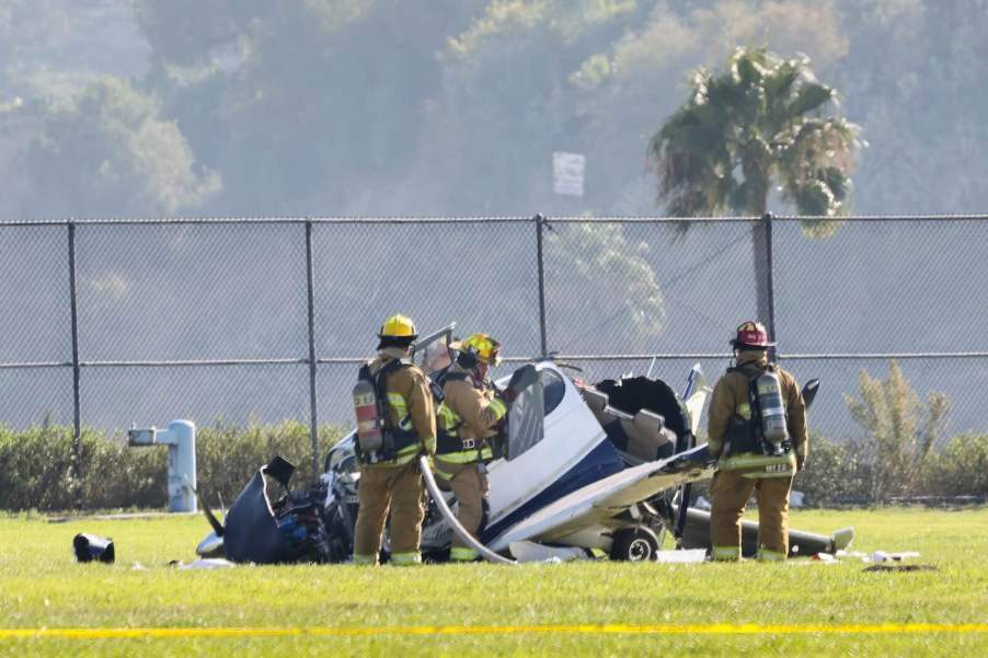 Fire fighters surround a small, ultralight airplane crash on a field, palm trees visible in the background.