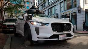 The grille of a Waymo self-driving taxi cab on a city street, buildings and trees visible in the background.