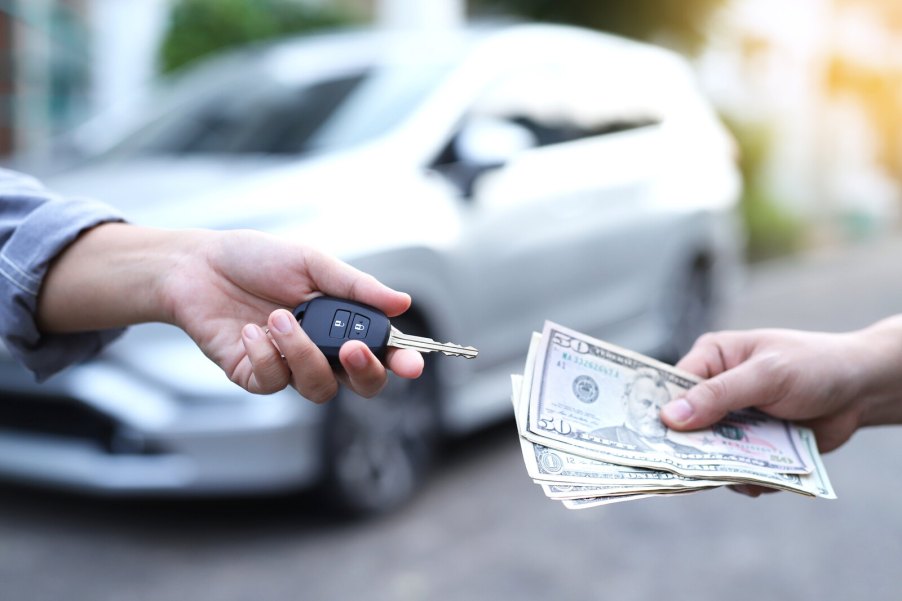 Hands exchanging car keys and cash with silver car parked in blurred background