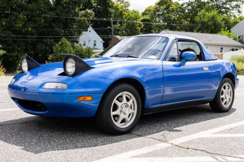 A blue 1994 Mazda MX-5 Miata parked with headlights open in low left front angle view
