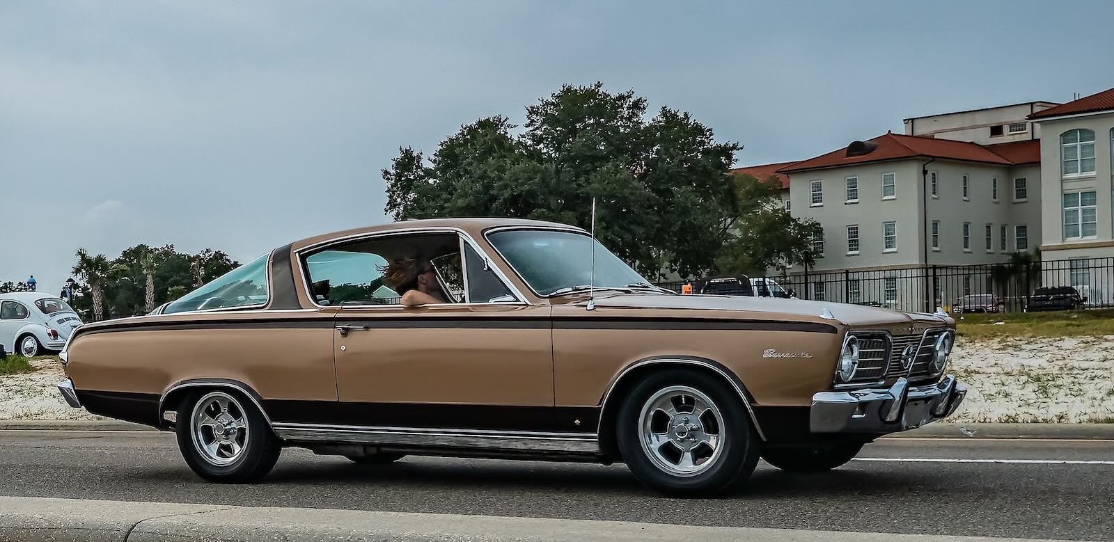 Tan Plymouth Barracuda muscle car driving on a road with buildings in the background.