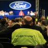 Factory workers look at a Ford sign on a stage during a meeting.
