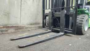The forks of a green forklift sitting on the pavement.