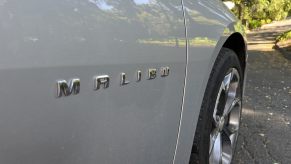 The Malibu badge on the door of a silver Chevy sedan, trees visible in the background.