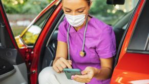 Blonde nurse wearing purple scrubs sits in a red car, on her phone