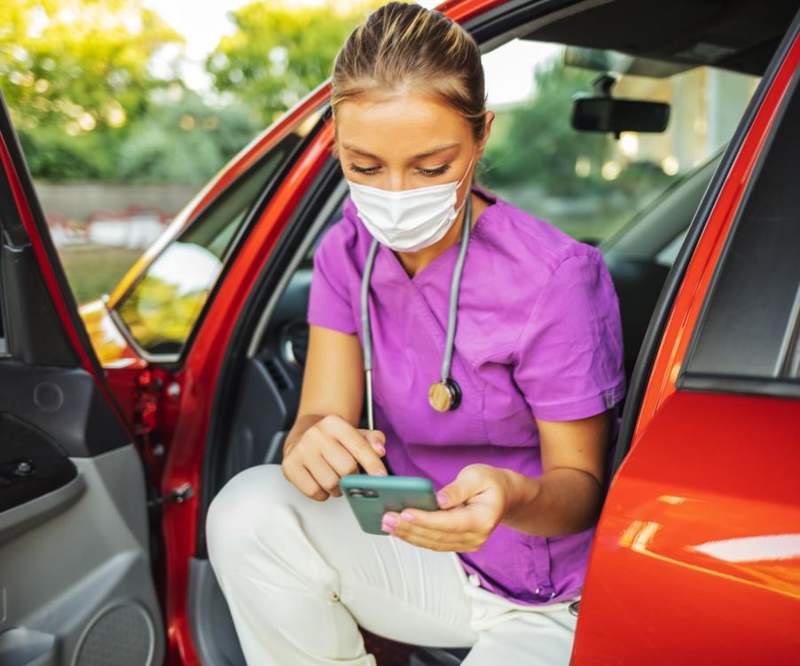 Blonde nurse wearing purple scrubs sits in a red car, on her phone