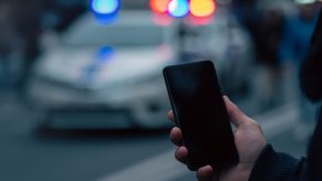 Hands hold a cell phone in front of a blurred police cruiser at dusk