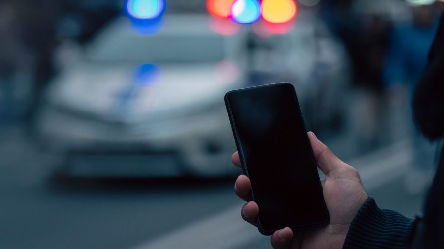 Hands hold a cell phone in front of a blurred police cruiser at dusk