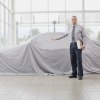 A car dealership salesman holding his hand out in front of a car covered in a white cloth