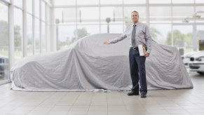 A car dealership salesman holding his hand out in front of a car covered in a white cloth
