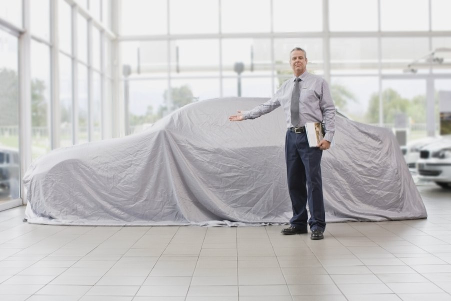 A car dealership salesman holding his hand out in front of a car covered in a white cloth