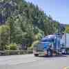 A semi-truck with a blue cab drives down a wooded highway
