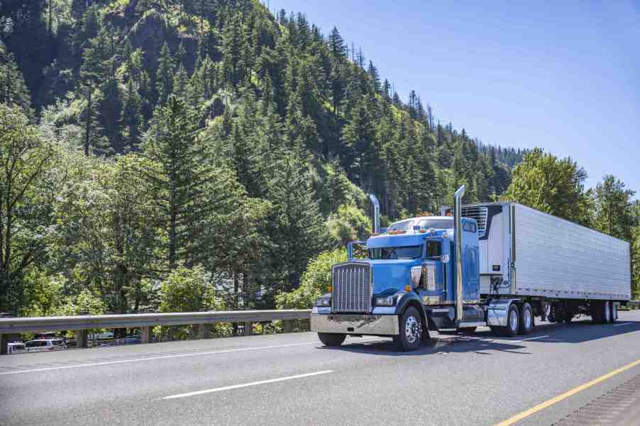 A semi-truck with a blue cab drives down a wooded highway