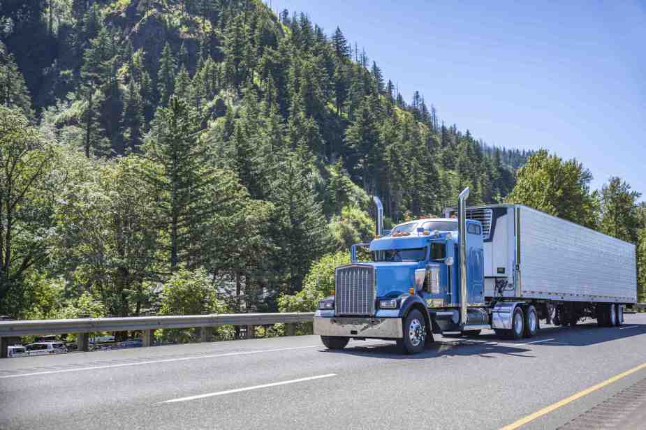 A semi-truck with a blue cab drives down a wooded highway