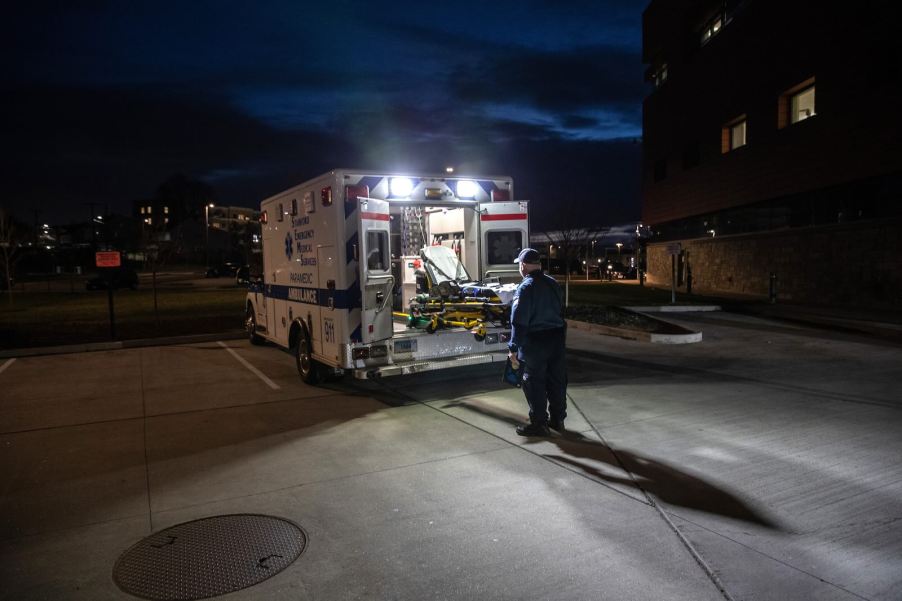 Man stands behind an open, empty ambulance in a parking lot at night.