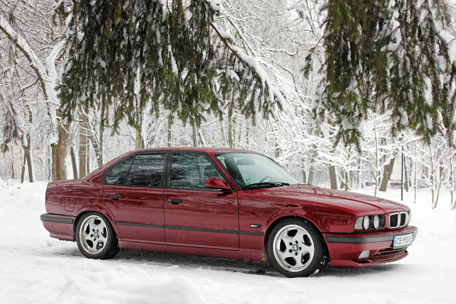 Red BMW M5 sports sedan parked in the woods, snow visible in the background.
