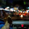 Driver holds the steering while and a beer, a city street visible in the background.