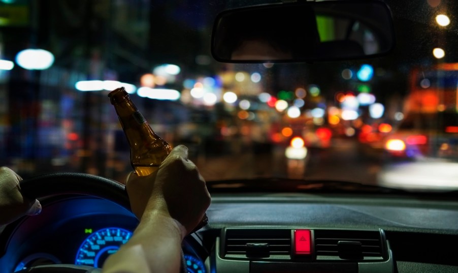 Driver holds the steering while and a beer, a city street visible in the background.