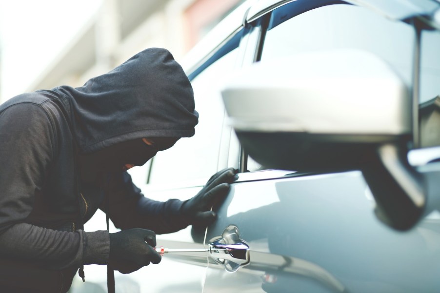 Thief in a hood and mask uses a screwdriver to get into a silver sedan