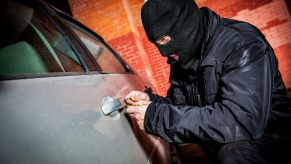 Thief in mask breaks into a silver car, a brick wall visible in the background.