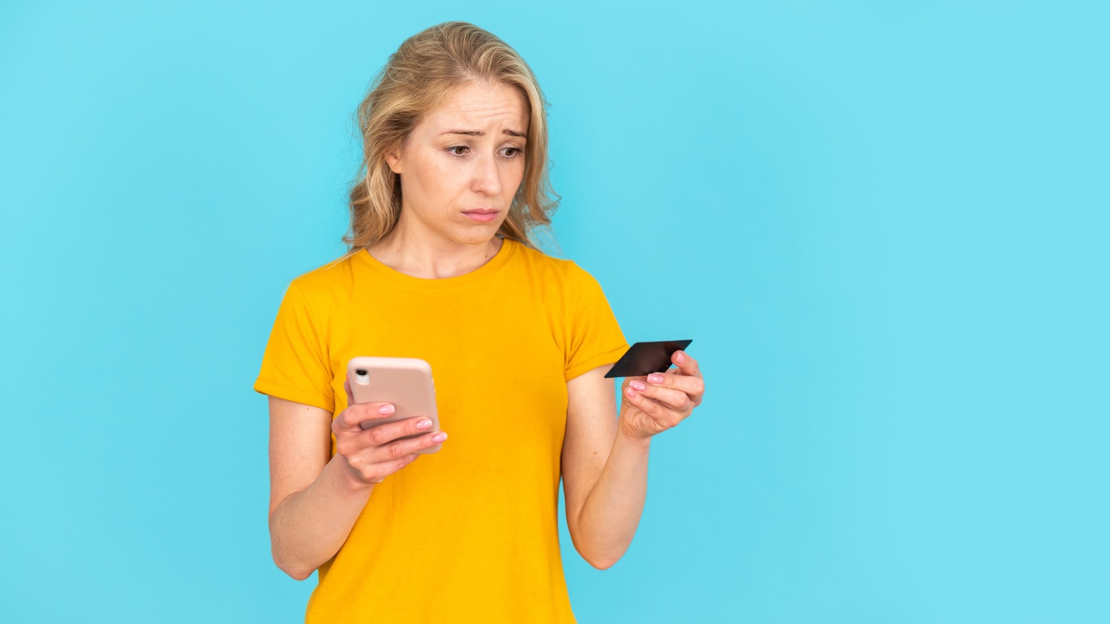 Sad woman in yellow shirt looks at her credit card and her phone's banking app.