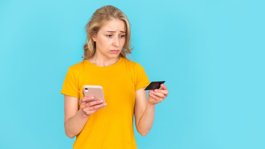 Sad woman in yellow shirt looks at her credit card and her phone's banking app.