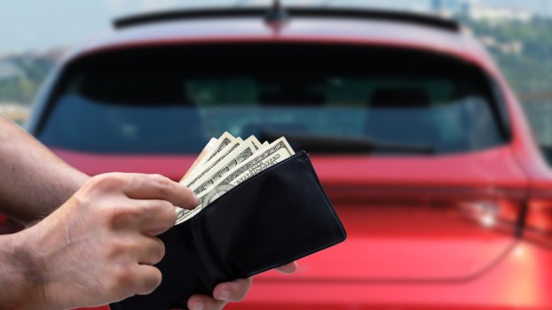 Car buyer pulls out cash, a red crossover SUV in the background.