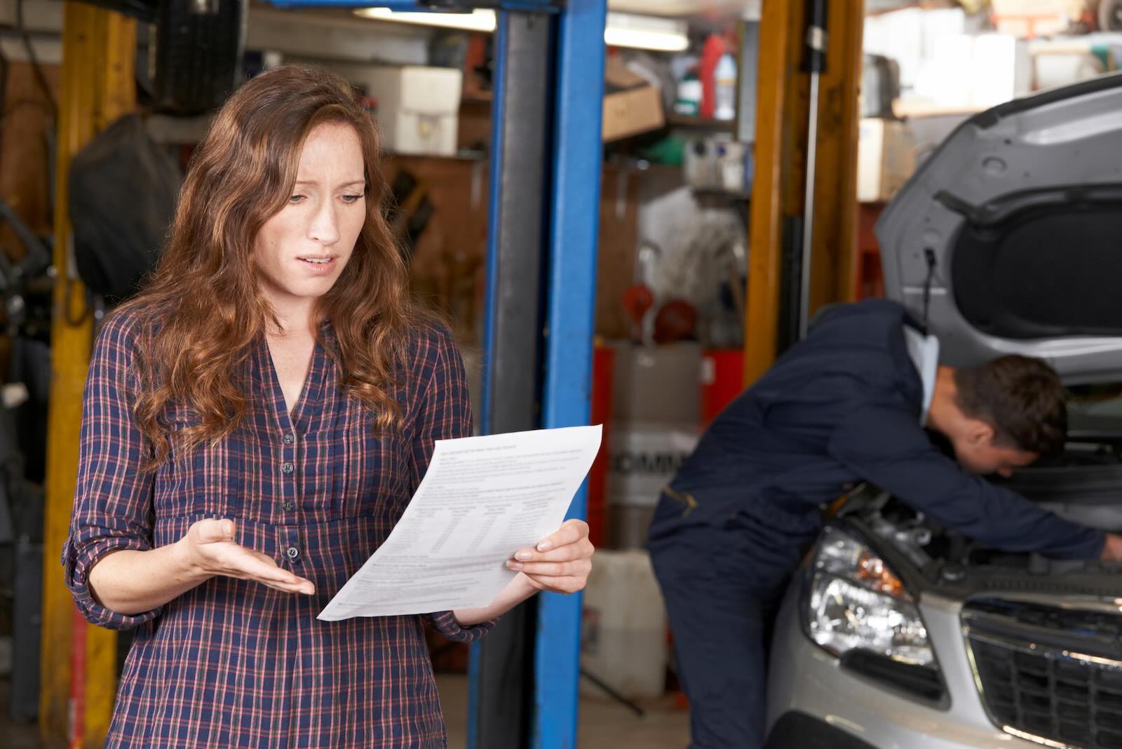 Woman holds Lexus dealership repair estimate in shock while mechanic works in the background.
