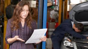 Woman holds Lexus dealership repair estimate in shock while mechanic works in the background.